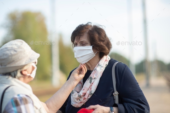 one senior woman helps another put on a medical face mask during a pandemic