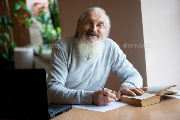 Bearded smiling old man with laptop and book sitting at table and write notes in library
