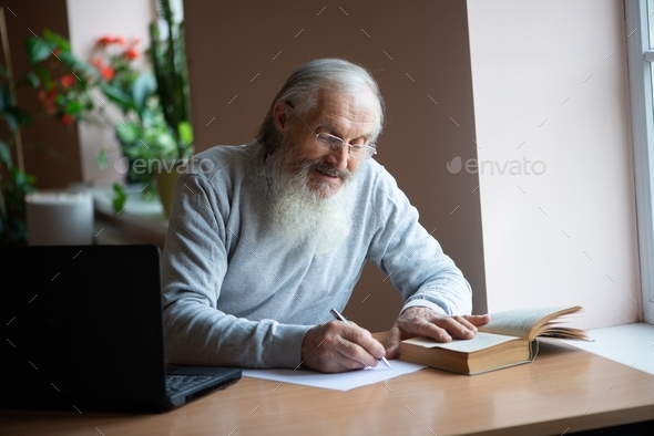 Bearded old man with laptop and book sitting at table and write notes in library