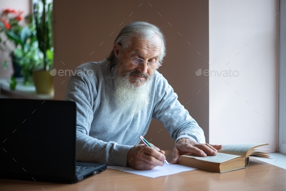 Bearded old man with laptop and book sitting at table and write notes in library