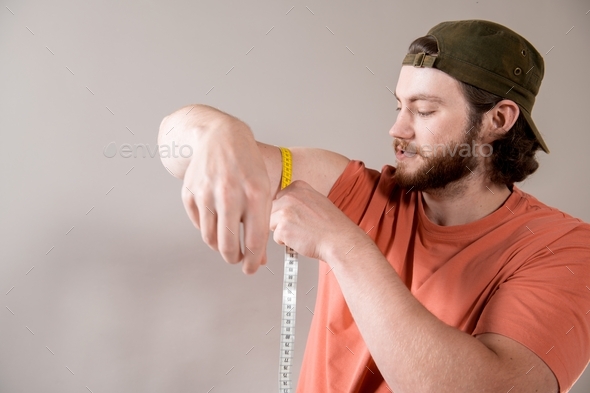 Bearded young caucasian man measuring biceps, muscles of his arm with a yellow tape measure.