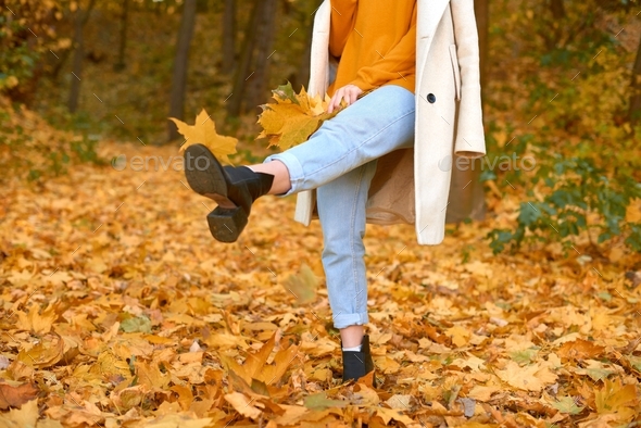 woman wearing stylish coat and orange pullover kicks fallen maple leaves and fool in autumn park