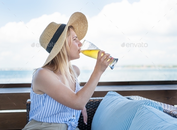 Attractive young blond woman in straw hat relaxing on a terrace drinking beer in bar on seaside