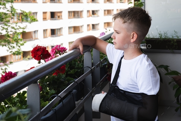 Sad boy with a broken arm in a cast looks sadly into yard from the balcony