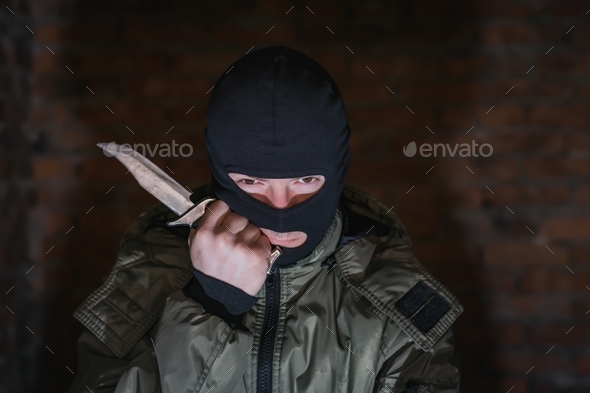 Tired bandit in balaclava and holding a knife on a brick wall background