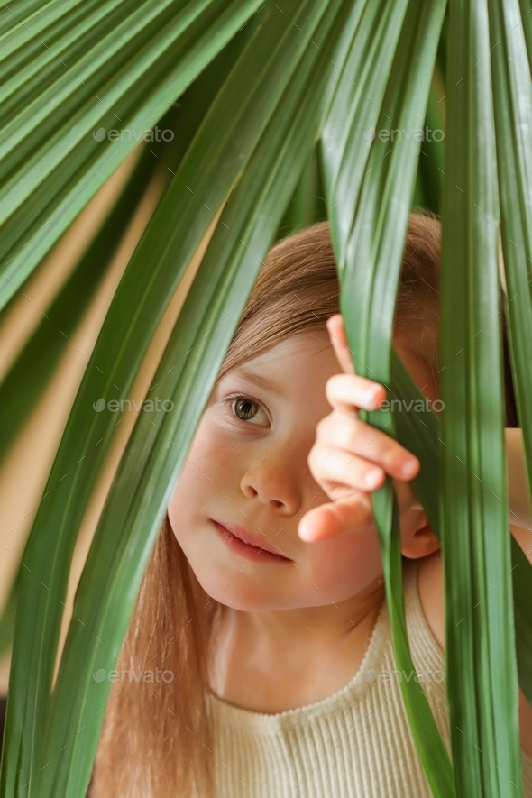 Little girl posing against the backdrop of a palm tree growing at home. Jungle-inspired aesthetics.