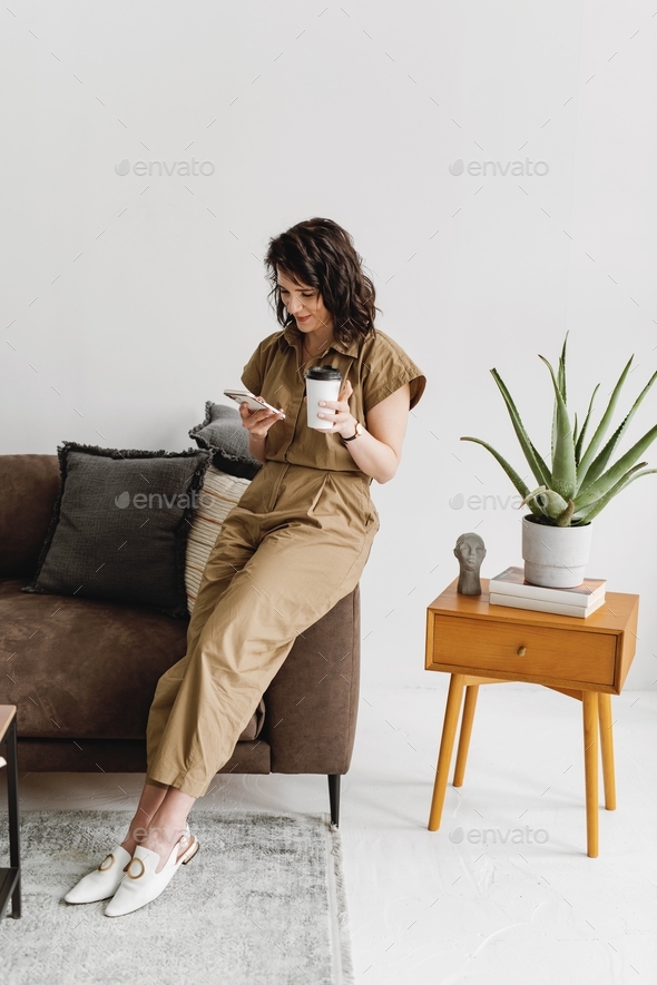Woman sitting on the edge of couch with coffee and using smartphone.
