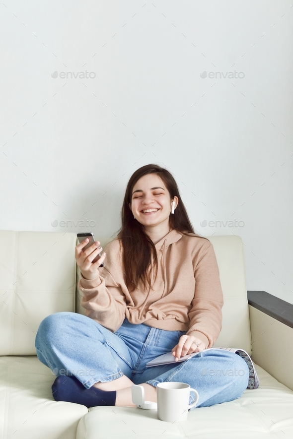 A young woman sits on the couch looks at the phone and laughs, wireless headphones in her ears.