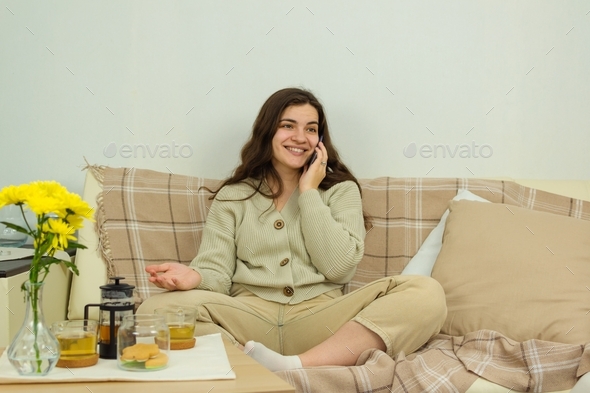 A young woman is sitting on a sofa, talking on a mobile phone, inviting friends and family to tea.