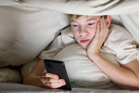 Boy under blanket at night in bed communicates on Internet. Child gadget addiction and insomnia