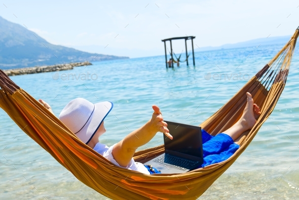 Work from anywhere. Young smiling woman, female freelancer in straw hat working on laptop on beach.