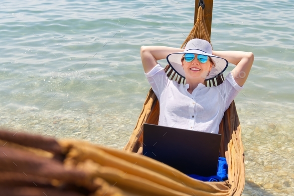 Work from anywhere. Young smilyng woman, female freelancer in straw hat working on laptop on beach.