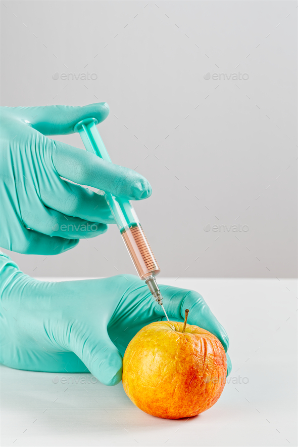 A hand in a medical glove inserts a syringe into apple. Harmful food