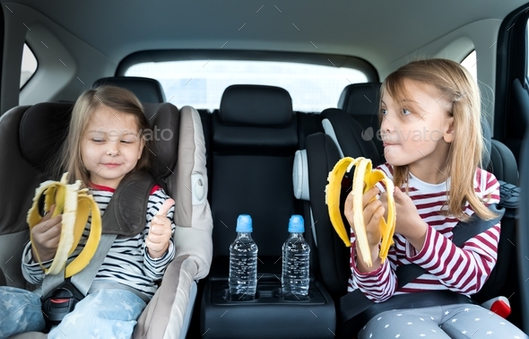 Little girls, sisters are driving in car, eating banana. Traveling on road in safe baby seats with c