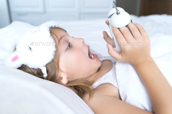 Little screaming girl sleeps in bed with big alarm,clock. Schoolgirl overslept, woke up late to stud