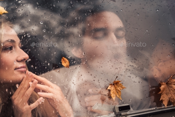 Young happy lovers couple behind wet misted window,rain drops.Drawing heart with finger.Autumn atmos