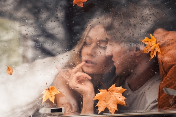 Young happy lovers couple behind wet misted window,rain drops.Drawing heart with finger.Autumn atmos