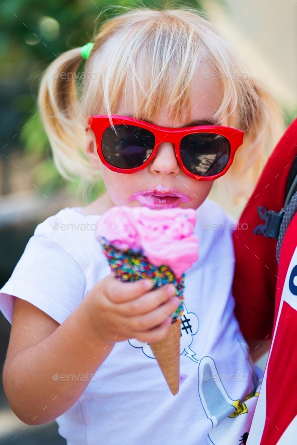 Cute little girl with messy face in red sunglasses eating melting ice cream cone