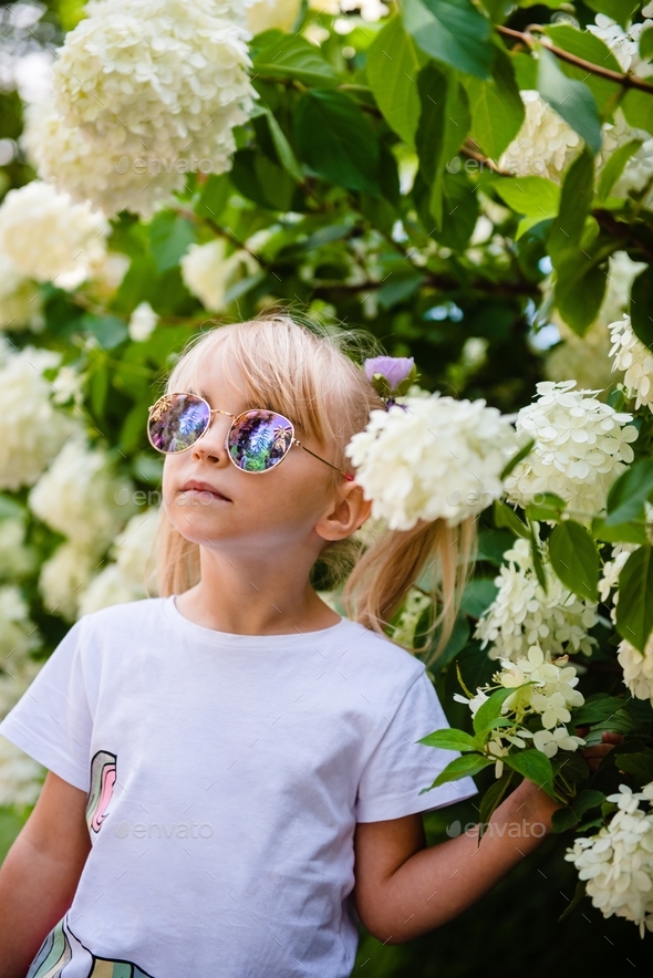 Beautiful little girl in stylish sunglasses in blooming flowers. - Stock Photo - Images