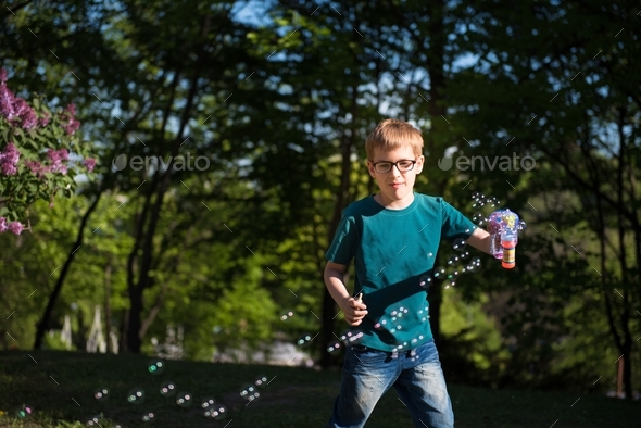 Boy in eyeglasses using soap bubbles gun to spread bubbles.