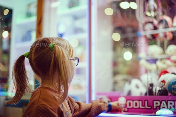 Little girl in eyeglasses playing a slot machine and trying to grab and win stuffed toys.