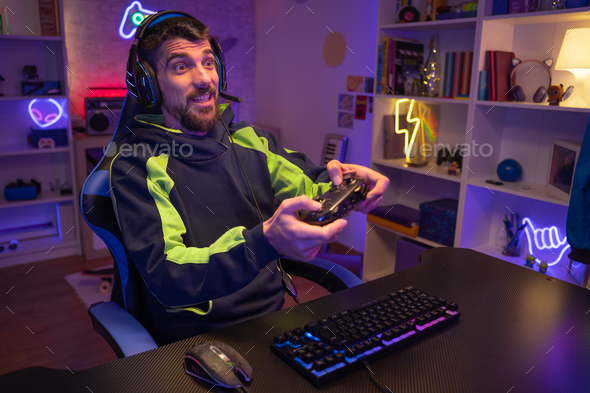 Happy guy gamer wear headphone play video game colorful neon lights computer in living room.