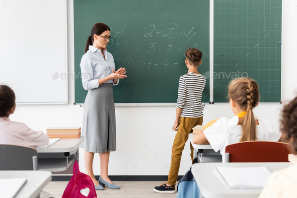 back view of schoolboy solving equations on chalkboard during math lesson near teacher and