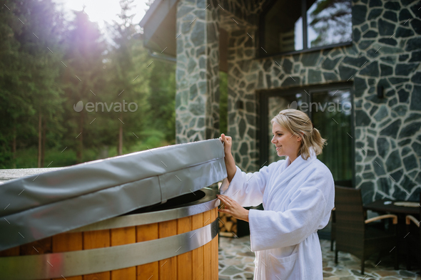 Woman in bathrobe opening lid of hot tub, checking temperature, ready for home spa procedure in hot