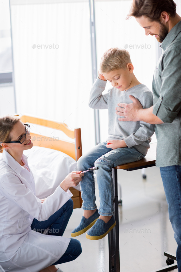 Female doctor with reflex hammer inspecting little boy sitting near father