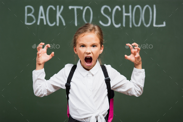selective focus of angry schoolgirl showing scaring gesture near chalkboard with back to school
