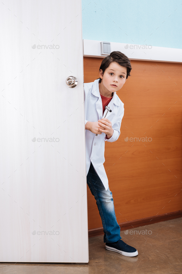 Scared boy doctor with reflex hammer standing in doorway and looking at camera
