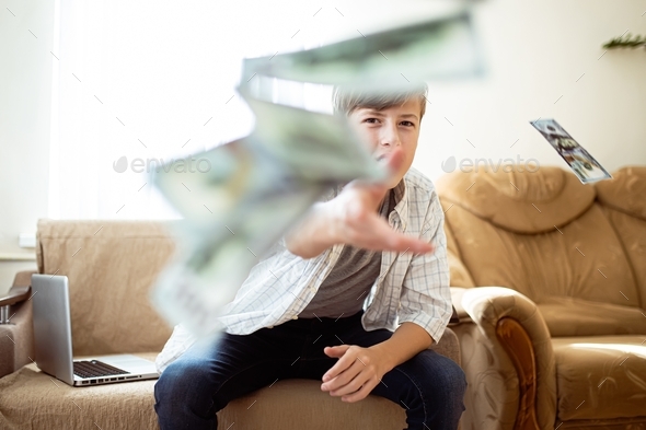 Boy flush with money. Boy throwing money around the room while sitting with laptop by the window