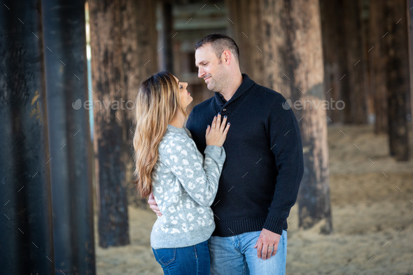 A couple dressed in sweaters standing under a pier romantically looking into each other eyes.  - Stock Photo - Images