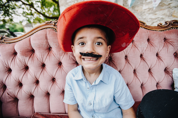Boy wearing a red Mexican hat and fake mustache sitting on a couch outside