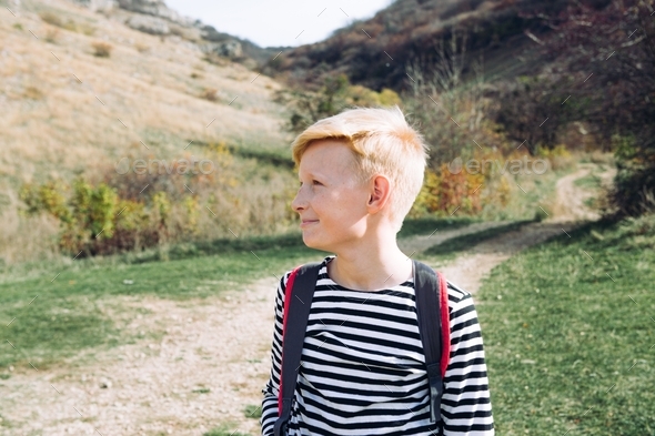A blond boy 10 years old in a striped jacket with a red backpack goes on a hike in the fall. Beautif