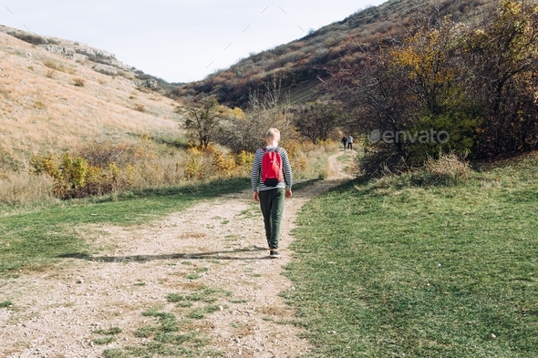 A blond boy 10 years old in a striped jacket with a red backpack goes on a hike in the fall.