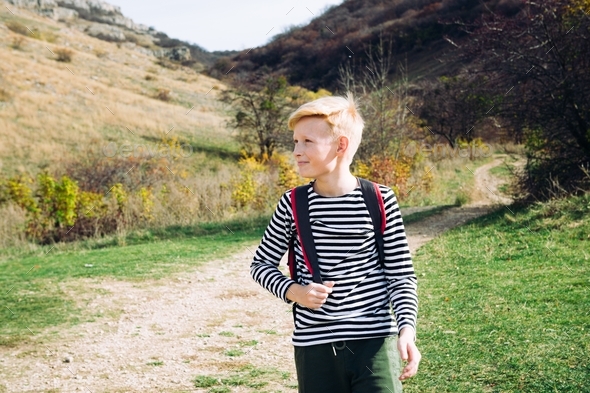 A blond boy 10 years old in a striped jacket with a red backpack goes on a hike in the fall.