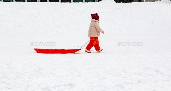 Little girl in the snow wearing orange snow pants carrying her orange sled behind her