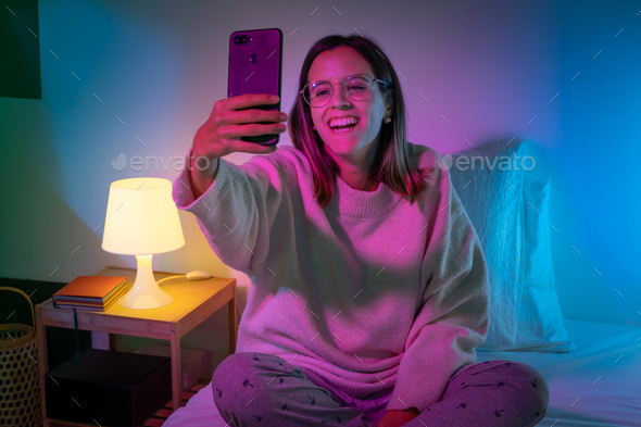 Beautiful young girl taking selfie with her phone on bed with neon colors room - Stock Photo - Images