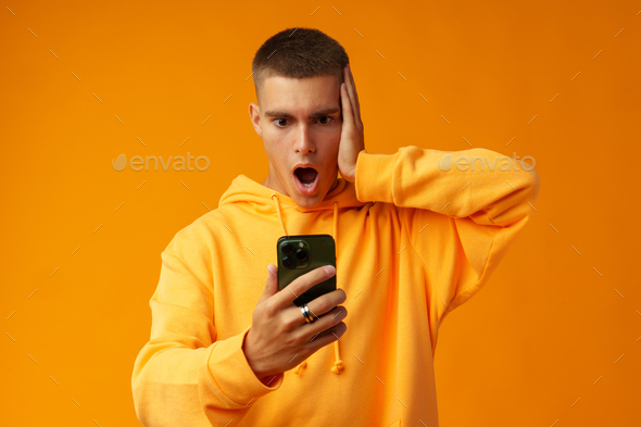 Young man expressing negativity while reading message on the mobile phone on yellow background
