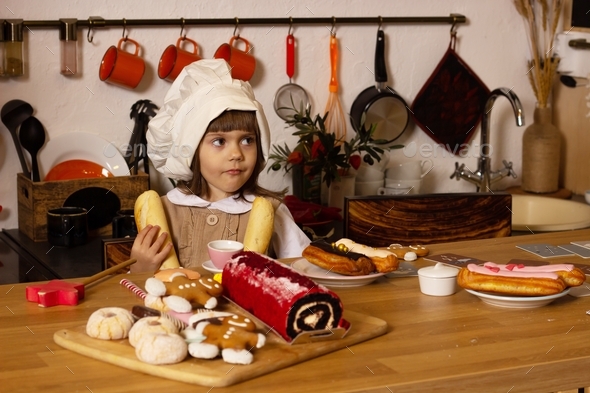 A girl in a chef's hat is sitting in the kitchen with baguettes, cakes, cake and gingerbread.