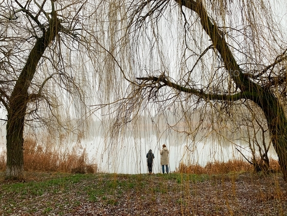 Two tiny figures of people standing under the trees at the foggy lakeside in autumn