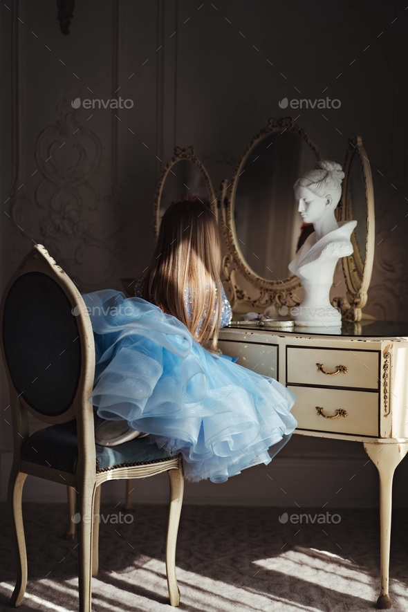 Pretty little girl in blue fancy dress sitting by dressing table looking in the mirror
