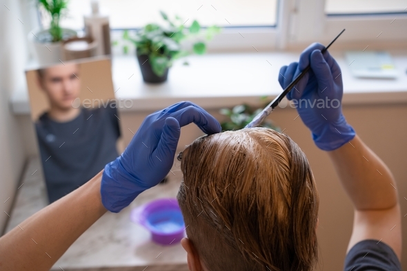 Young caucasian male with blue gloves applying bleaching dye on hairs in front of mirror at home