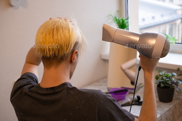 Young man sitting in front of mirror after bleaching his hair and drying his hair with a hairdryer