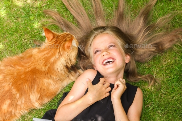 Beautiful girl with long hair laughing and lying on grass with maine coon cat sniffing her head