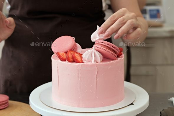 Woman pastry chef decorates pink cake with macaroons and berries, close-up. Cake making process