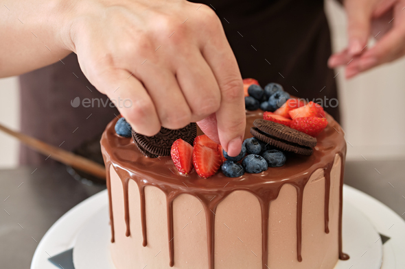 Woman pastry chef decorates chocolate cake with berries and cookies, close-up. Cake making process