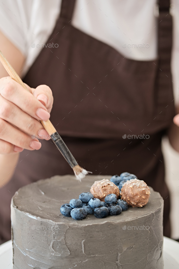 Woman pastry chef decorates grey cake with blueberries and glitter, close-up. Cake making process