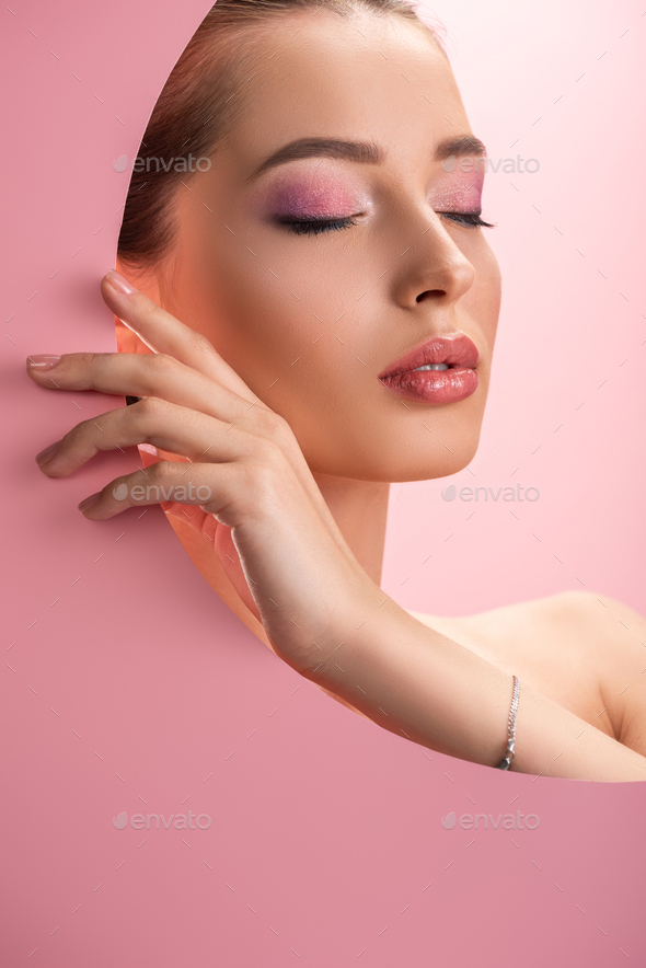 beautiful naked woman with shiny makeup in round paper hole with closed eyes isolated on pink